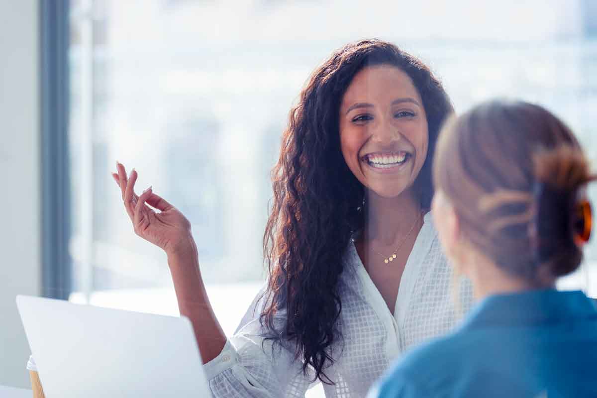 Business colleagues having a conversation. They are both young business people casually dressed in a modern office. Could be an interview or consultant working with a client. She is listening and smiling. One person has her back to us. Mixed ethnic group. One is African American and the other is Caucasian