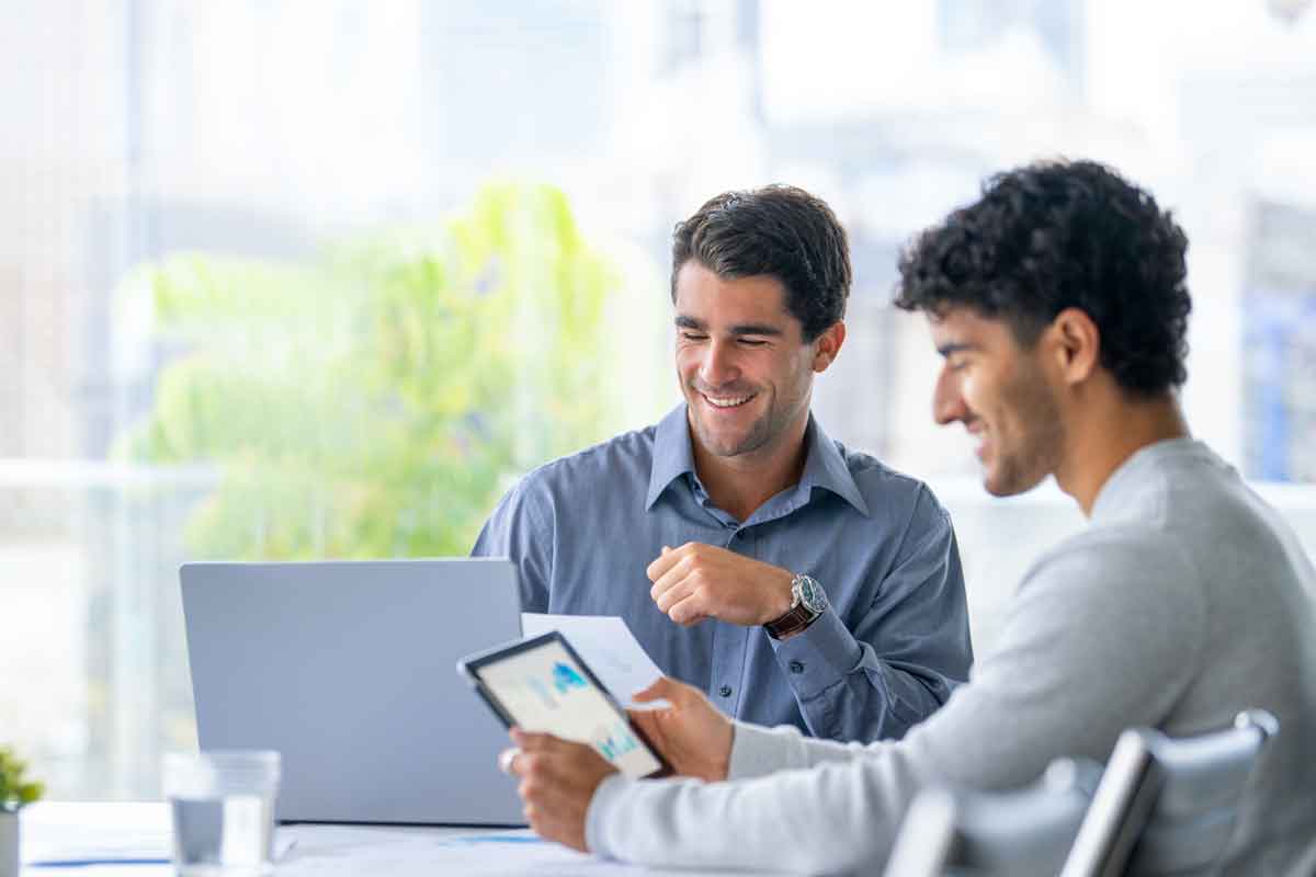 Two businessmen working on a digital tablet and laptop computer in the office. Finance Charts and graphs can be seen on the digital tablet