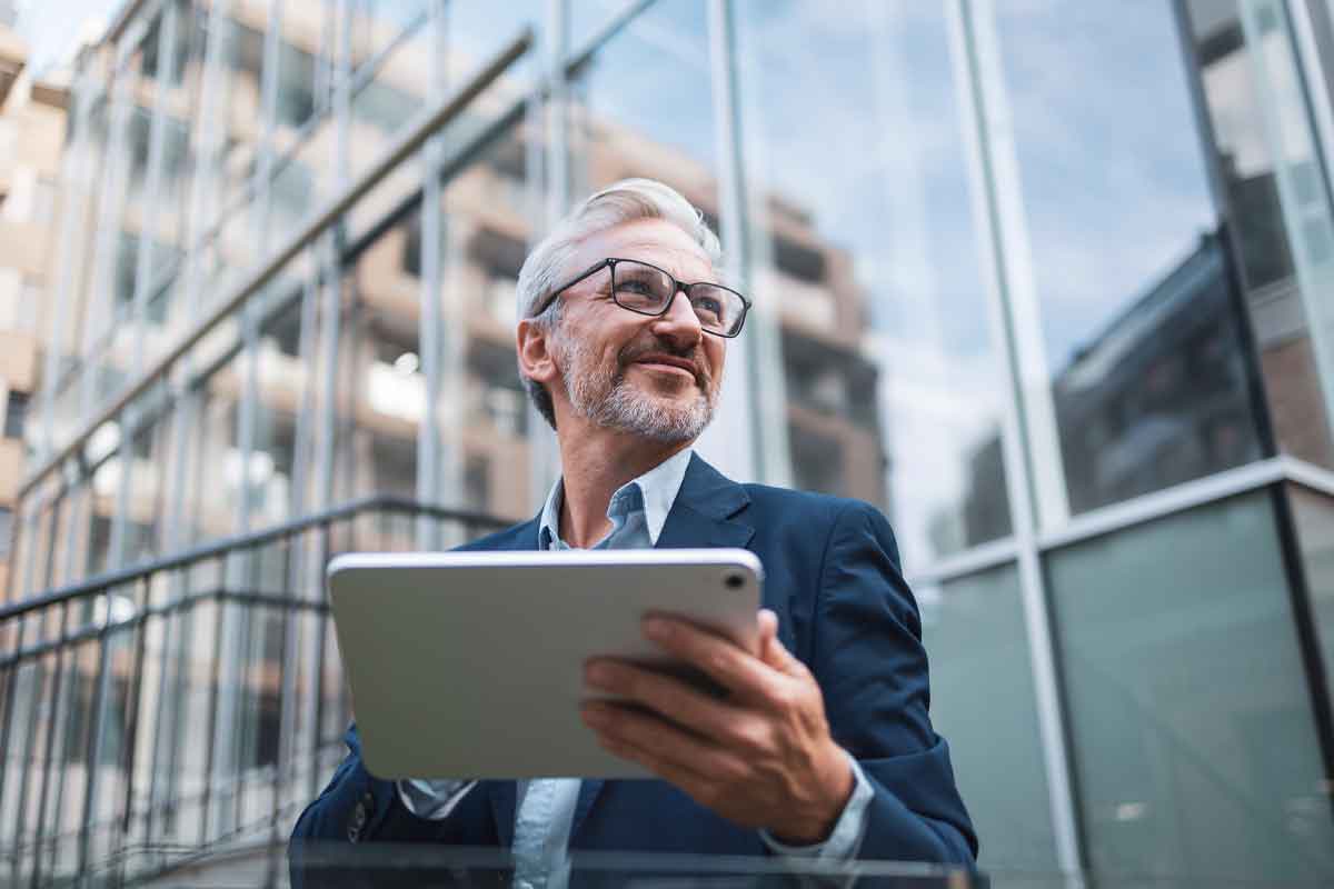 A mature businessman with glasses and gray hair stands confidently outside a modern glass building, holding a tablet. The image conveys professionalism, success, and a forward-thinking attitude.